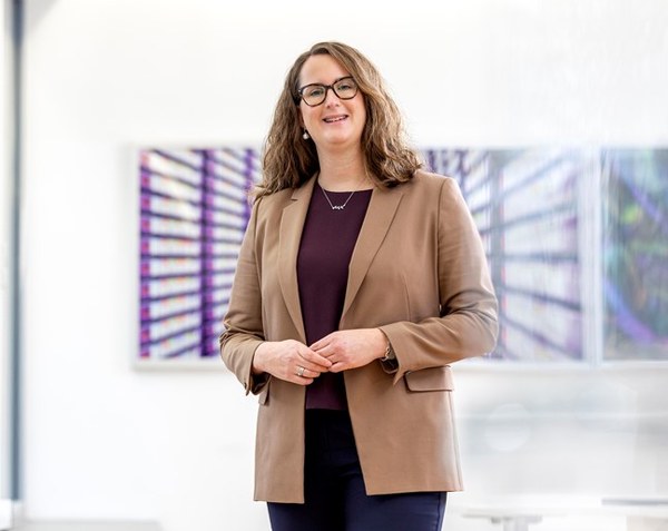 Dr. Sabrina Röttger-Wirtz steht im Zentrum des Bildes und lächelt in die Kamera. Im Hintergrund sieht man verschwommen ein Bild an einer weißen Wand.